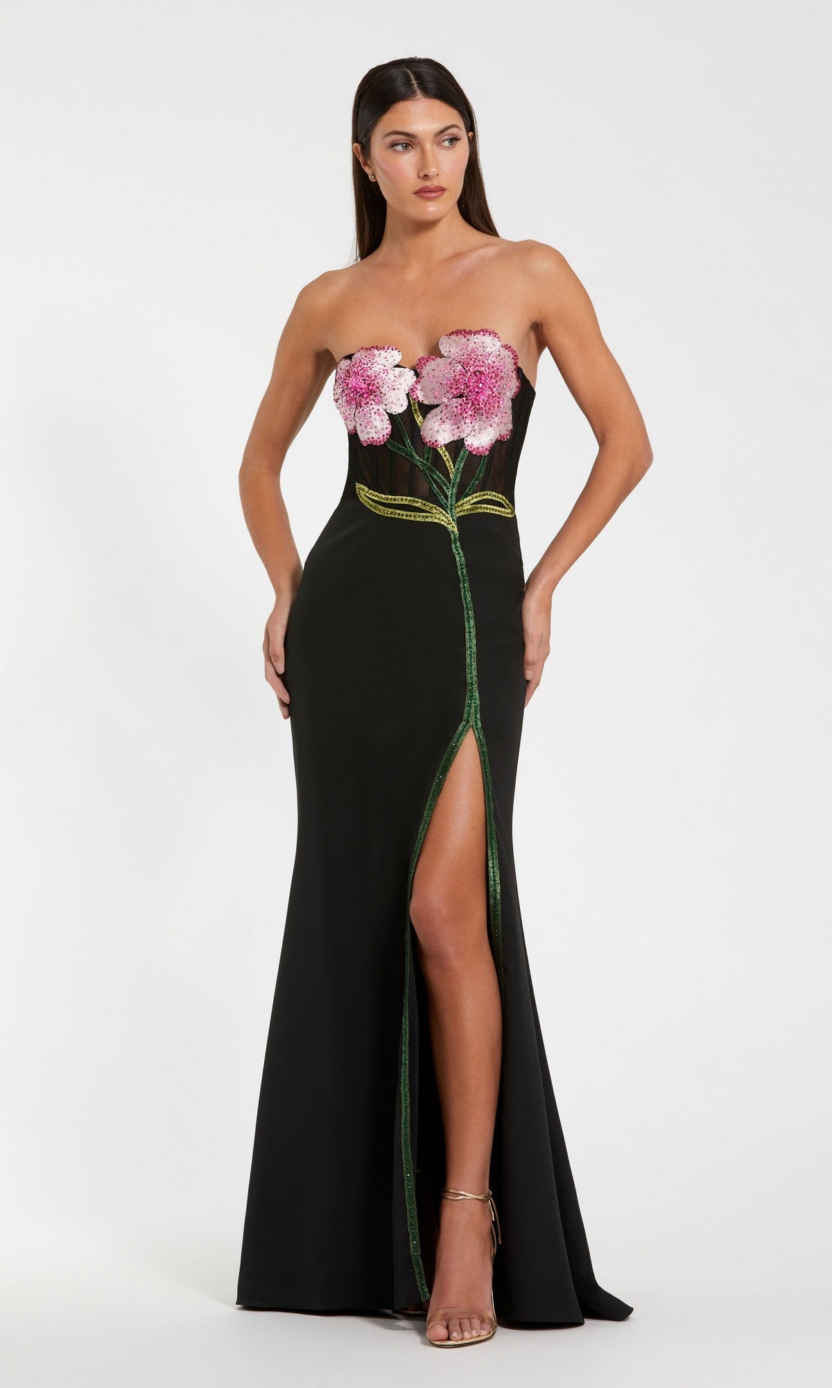 A woman wears the Mac Duggal 20682 long formal dress in black, featuring a strapless crepe design with pink flower appliqués on the bodice, a green stem down the front, and a high leg slit—ideal for special occasions.